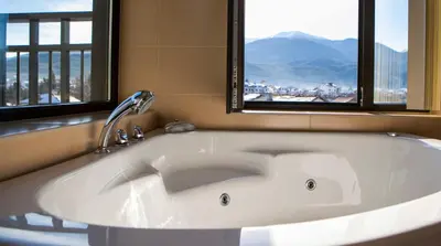 a large white bath tub in a bathroom with two windows at Къщи за гости КООП Добринище - Holiday Villages COOP Dobrinishte in Dobrinishte