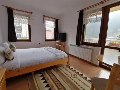 a bedroom with a bed and a table and windows at Къща за гости Камината in Trigrad